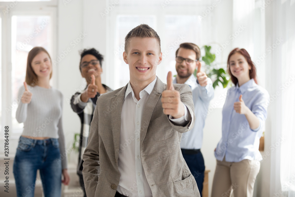 Smiling millennial male employee or worker stand foreground showing thumbs up looking at camera, diverse colleagues at back support with recommendation gesture, multiracial team recommend or approve Smiling millennial male employee or worker stand foreground showing thumbs up looking at camera, diverse colleagues at back support with recommendation gesture, multiracial team recommend or approve