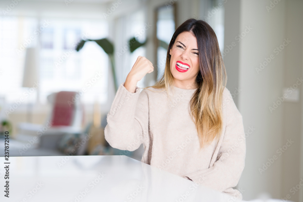 Young beautiful woman wearing winter sweater at home angry and mad raising fist frustrated and furious while shouting with anger. Rage and aggressive concept. Young beautiful woman wearing winter sweater at home angry and mad raising fist frustrated and furious while shouting with anger. Rage and aggressive concept.