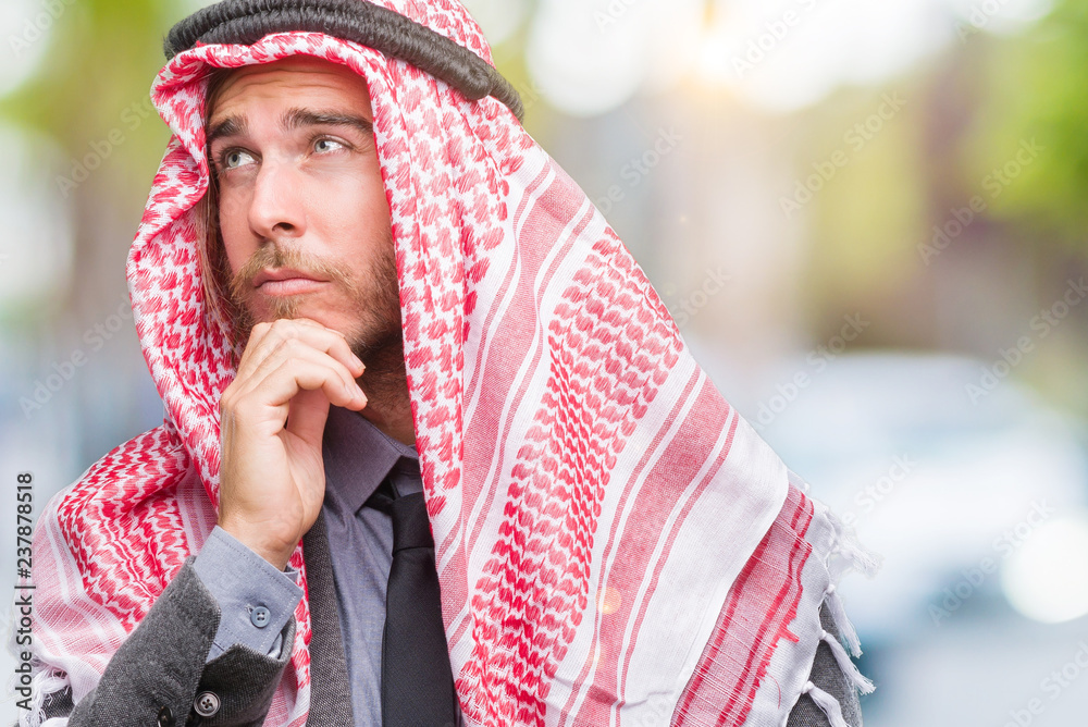 Young handsome arabian man with long hair wearing keffiyeh over isolated background with hand on chin thinking about question, pensive expression. Smiling with thoughtful face. Doubt concept.