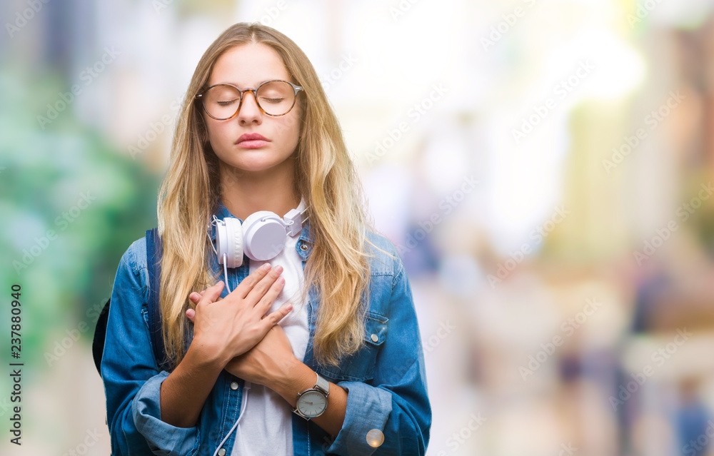 Young beautiful blonde student woman wearing headphones and glasses over isolated background smiling with hands on chest with closed eyes and grateful gesture on face. Health concept.