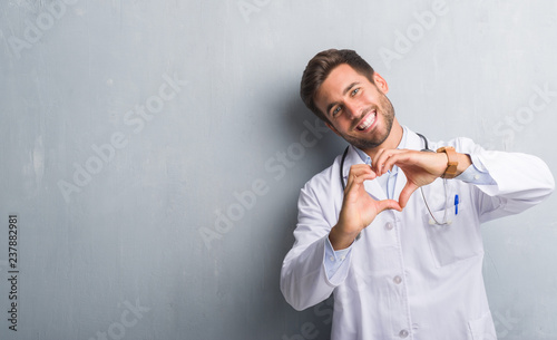 Handsome young doctor man over grey grunge wall smiling in love showing heart symbol and shape with hands. Romantic concept.