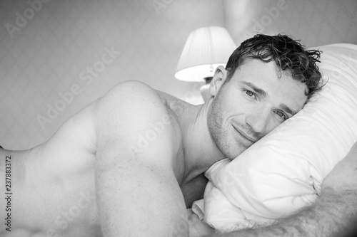 Fotografie Black and white picture of handsome naked man lying on hotel room bed sheets