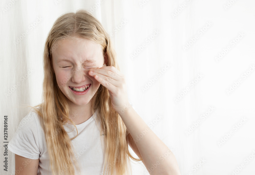 oung girl with curly and blonde hair trying on her mother's moisturizing patches from edema and tired eyes. Isolated photo on white background. Dust gets into girl's eyes. Free space copy for text oung girl with curly and blonde hair trying on her mother's moisturizing patches from edema and tired eyes. Isolated photo on white background. Dust gets into girl's eyes. Free space copy for text