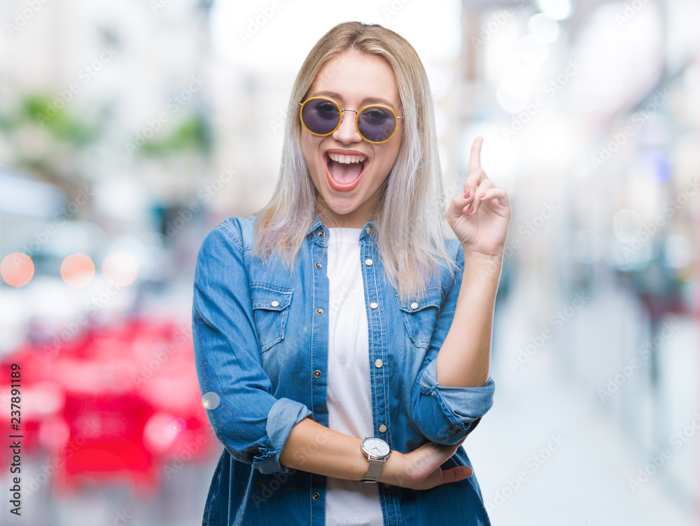 Young blonde woman wearing sunglasses over isolated background pointing finger up with successful idea. Exited and happy. Number one.