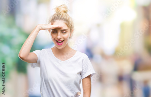 Young beautiful blonde woman wearing white t-shirt over isolated background very happy and smiling looking far away with hand over head. Searching concept.