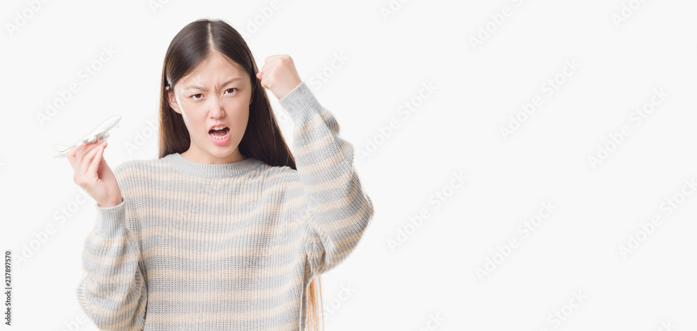 Young Chinese woman over isolated background holding aircraft toy annoyed and frustrated shouting with anger, crazy and yelling with raised hand, anger concept Young Chinese woman over isolated background holding aircraft toy annoyed and frustrated shouting with anger, crazy and yelling with raised hand, anger concept