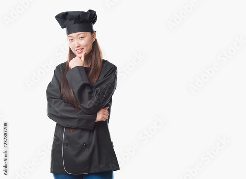 Young Chinese woman over isolated background wearing chef uniform looking confident at the camera with smile with crossed arms and hand raised on chin. Thinking positive.