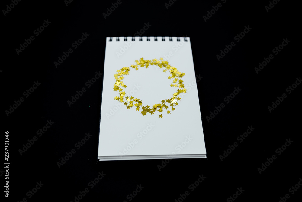 Obraz premium Spiral Notebook, realistic mockup White scetchbook on black abstract background with golden stars confetti.Horizontal top view..