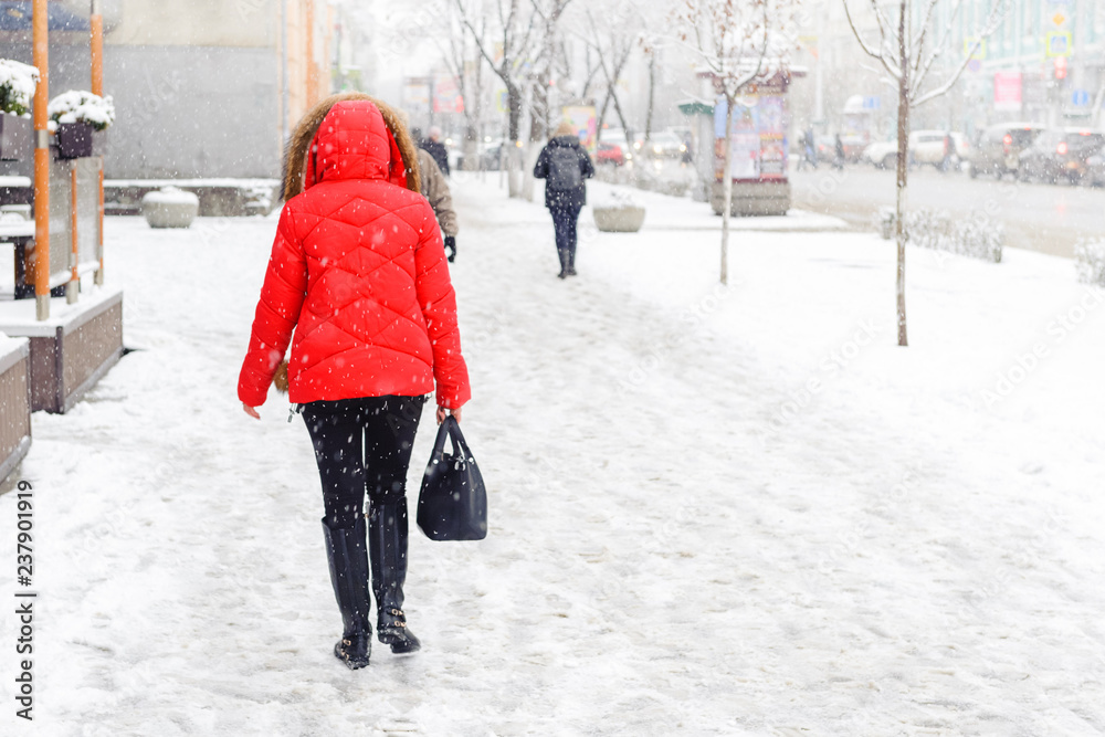 Winter, city, December, it is snowing, a woman in a red jacket walking down the street, rear view