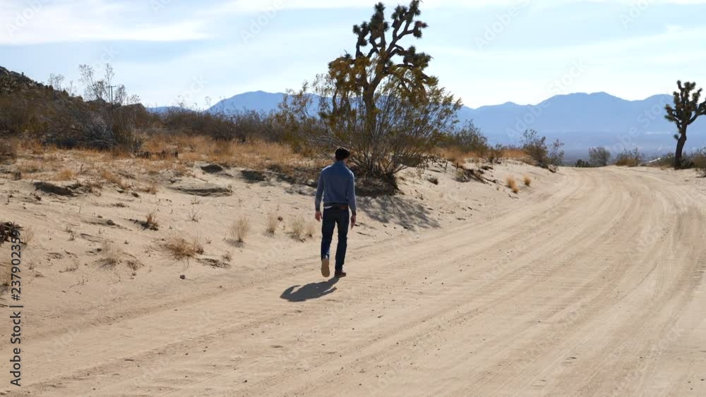 A man walking alone in slow motion down a hot dry desert road with Joshua trees and mountains in the landscape.