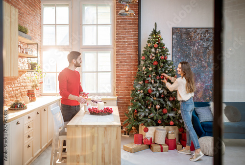 Young couple preparing for a New Year decorating their beautiful apartment standing in the dining room with Christmas tree on the background