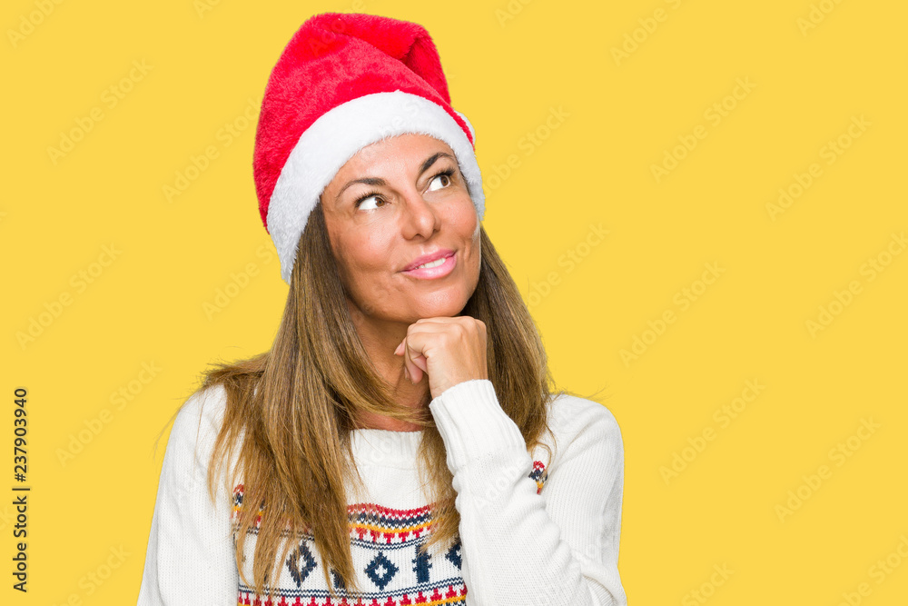 Middle age adult woman wearing winter sweater and chrismat hat over isolated background with hand on chin thinking about question, pensive expression. Smiling with thoughtful face. Doubt concept. Middle age adult woman wearing winter sweater and chrismat hat over isolated background with hand on chin thinking about question, pensive expression. Smiling with thoughtful face. Doubt concept.