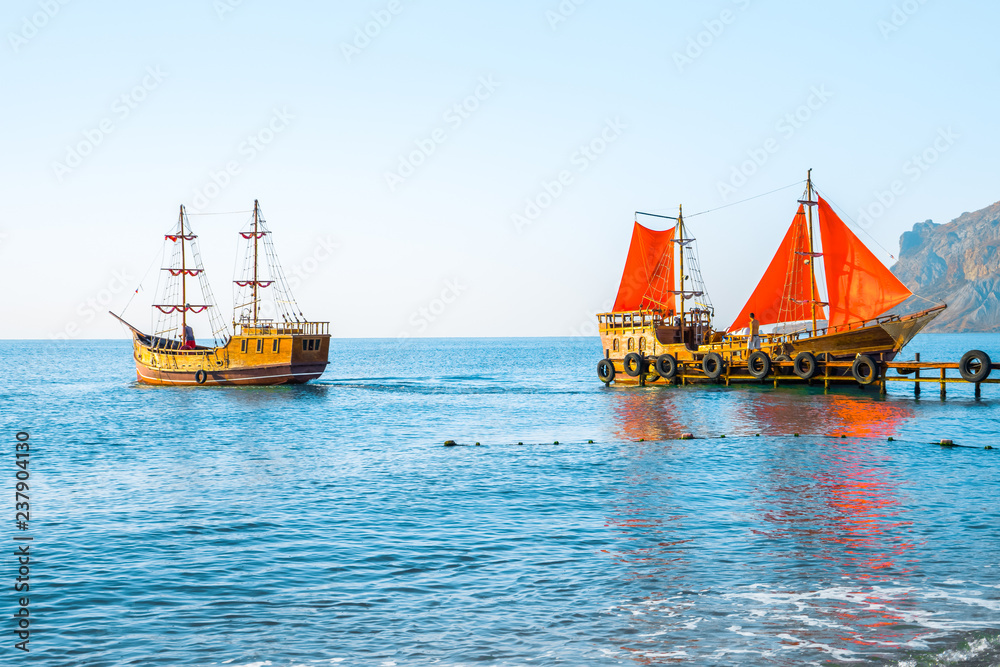 Obraz premium Two old wooden ships. One of them with red sails on the pier.