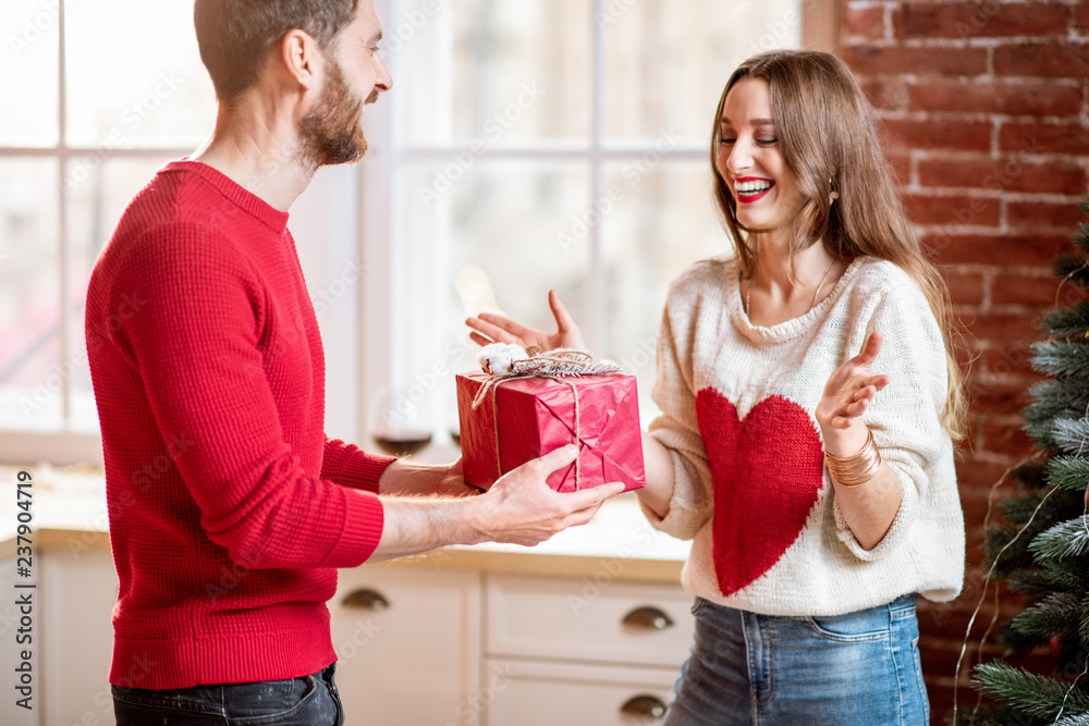 Man giving a New Year gift for a young excited woman standing near the Christmas tree at home Man giving a New Year gift for a young excited woman standing near the Christmas tree at home