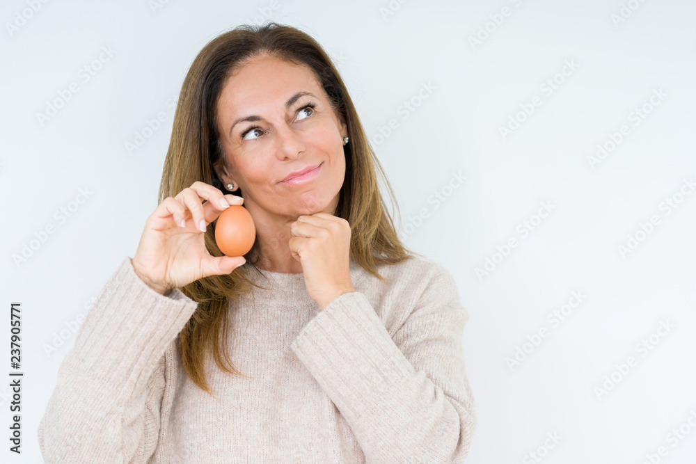 Middle age woman holding fresh egg over isolated background serious face thinking about question, very confused idea Middle age woman holding fresh egg over isolated background serious face thinking about question, very confused idea
