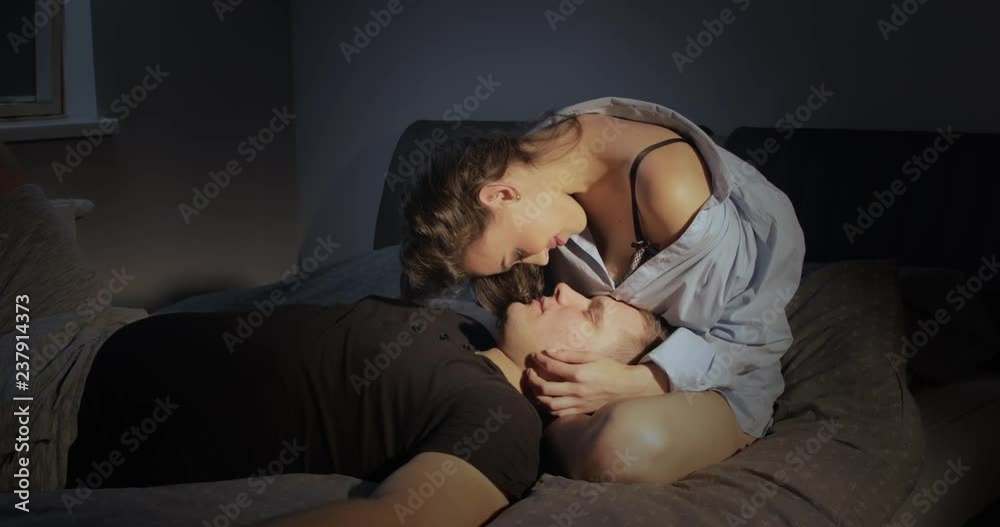 A young couple in love as the man lays in her lap in the bedroom and they lovingly gaze into each other's eyes as she gives him sweet kisses
