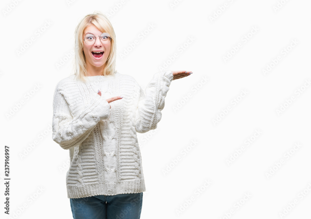 Young beautiful blonde woman wearing winter sweater and glasses over isolated background amazed and smiling to the camera while presenting with hand and pointing with finger. Young beautiful blonde woman wearing winter sweater and glasses over isolated background amazed and smiling to the camera while presenting with hand and pointing with finger.