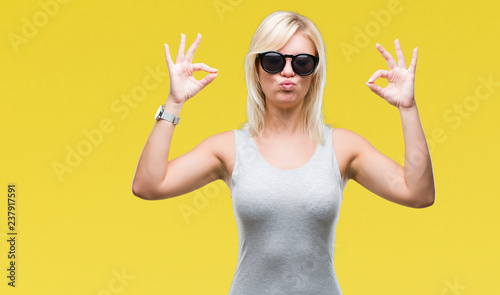Young beautiful blonde woman wearing sunglasses over isolated background relax and smiling with eyes closed doing meditation gesture with fingers. Yoga concept.