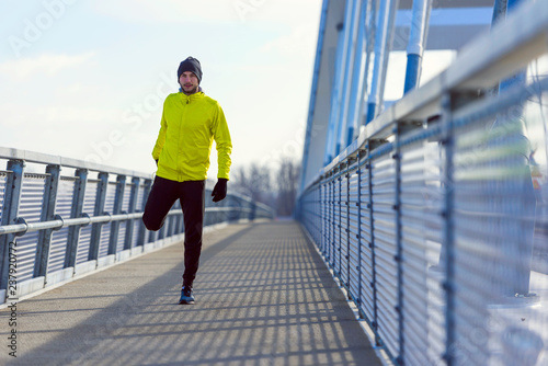 Young athlete man stretching his muscles before running on bridge on sunny winter day