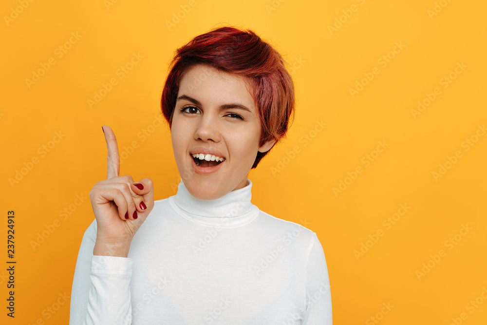 Woman portrait. Emotions. Ideas. Girl with short red hair is pointing up and winking at camera, on an orange background