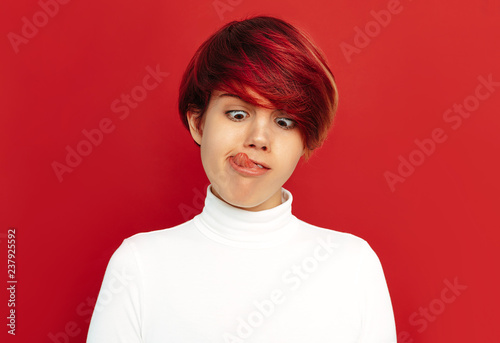 Woman portrait. Emotions. Girl with short red hair is making faces, on a red background