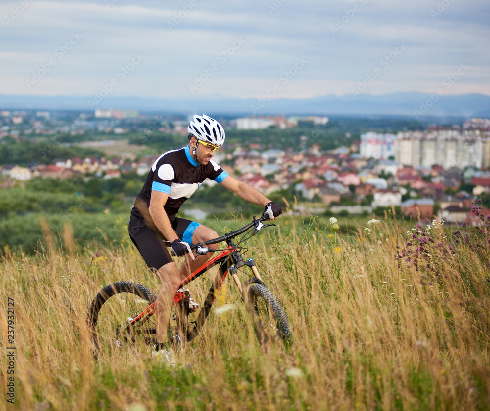 Side view of sporty man wearing sportswear and helmet riding bicycle on trail in grass. Concept of healthy lifestyle, well being, motivation and recreation. Urban background.