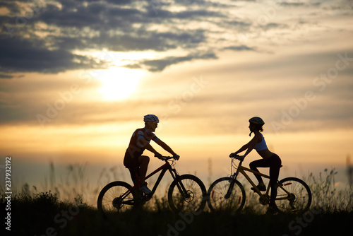 Two cyclists, woman and man standing opposite each other and posing with bikes. Silhouettes of couple, looking at each other, against amazing and wonderful background of sky with clouds and sunset.