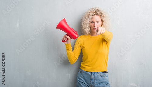 Young blonde woman over grunge grey background holding megaphone pointing with finger to the camera and to you, hand sign, positive and confident gesture from the front
