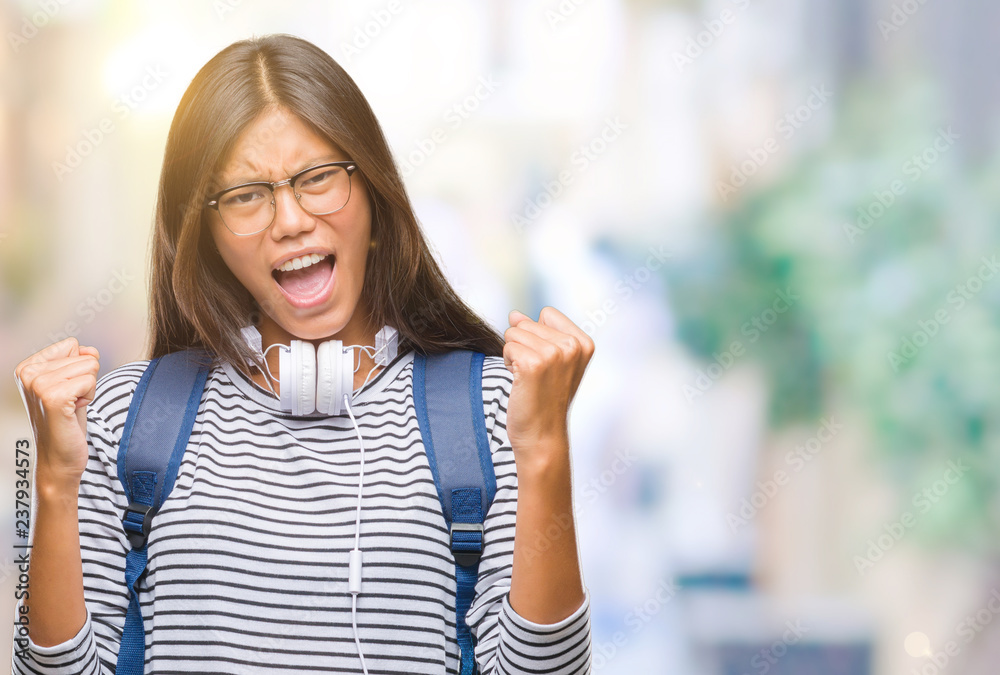 Young asian student woman wearing headphones and backpack over isolated background very happy and excited doing winner gesture with arms raised, smiling and screaming for success. Celebration concept.