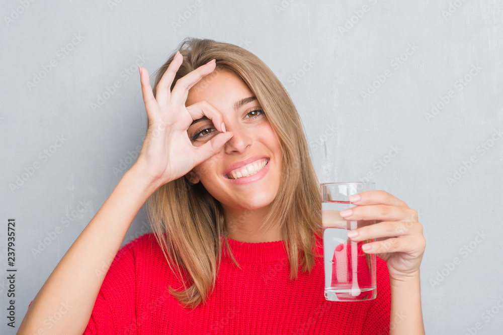 Beautiful young woman over grunge grey wall drinking a glass of water with happy face smiling doing ok sign with hand on eye looking through fingers Beautiful young woman over grunge grey wall drinking a glass of water with happy face smiling doing ok sign with hand on eye looking through fingers