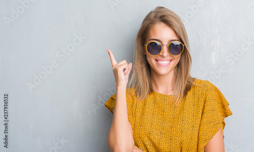 Beautiful young woman standing over grunge grey wall wearing retro sunglasses surprised with an idea or question pointing finger with happy face, number one