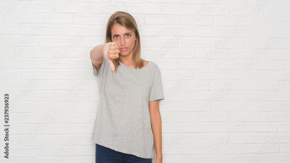 Beautiful young woman over white brick wall looking unhappy and angry showing rejection and negative with thumbs down gesture. Bad expression. Beautiful young woman over white brick wall looking unhappy and angry showing rejection and negative with thumbs down gesture. Bad expression.