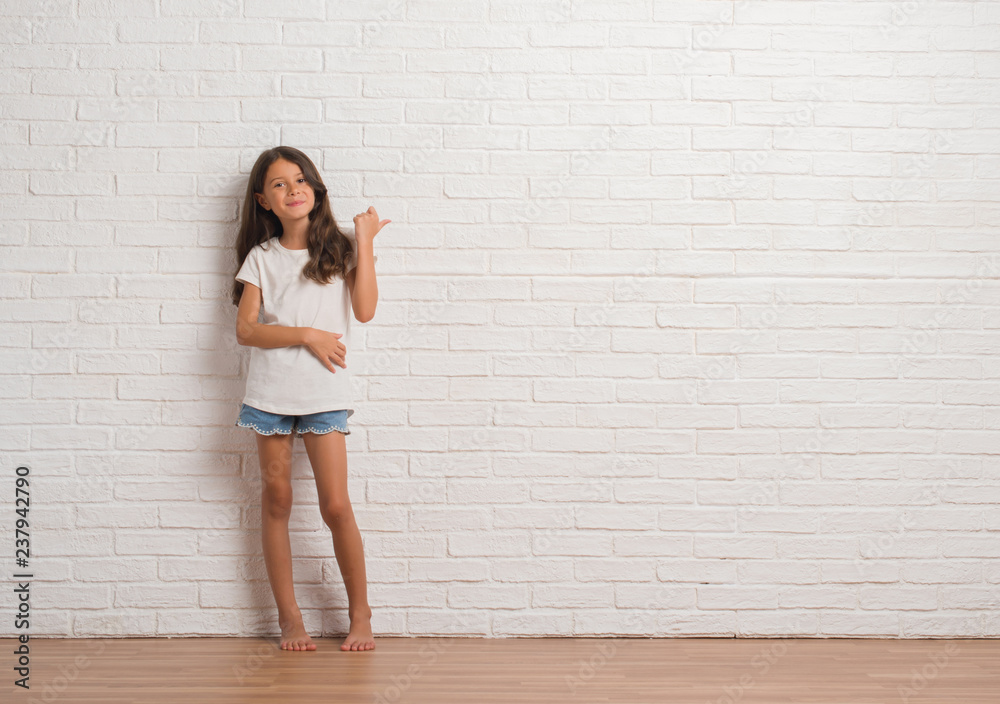 Young hispanic kid stading over white brick wall smiling with happy face looking and pointing to the side with thumb up. Young hispanic kid stading over white brick wall smiling with happy face looking and pointing to the side with thumb up.