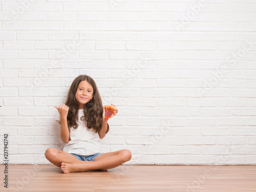 Young hispanic kid sitting on the floor over white brick wall eating pizza slice pointing and showing with thumb up to the side with happy face smiling