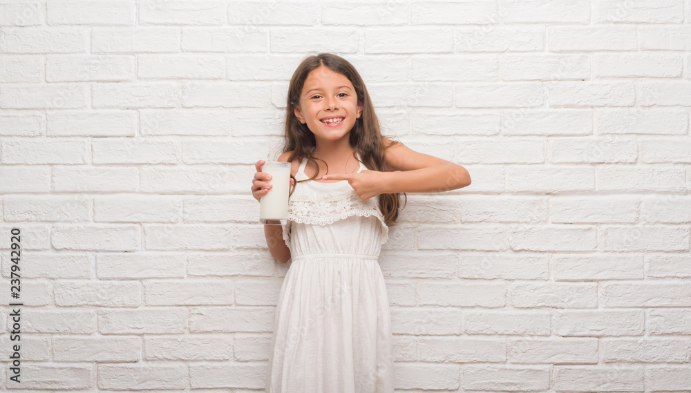 Young hispanic kid over white brick wall drinking a glass of milk very happy pointing with hand and finger