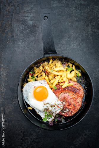 Traditional fried Allgauer veal roast roll sausage slices with fried egg in a cast-iron skillet with spices as closeup on a black board