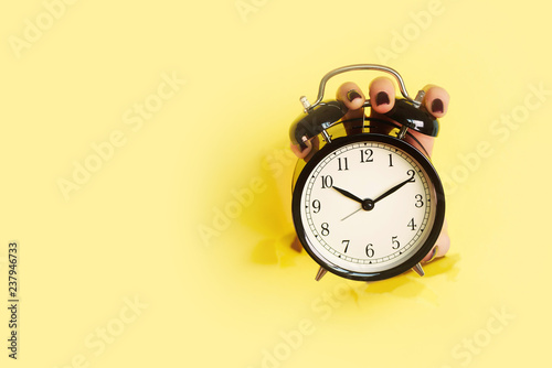 Hand holding black alarm clock through hole in yellow paper background. Wake up alert concept. Morning routine. Banner with copy space