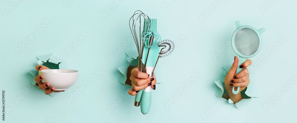 Woman hand holding kitchen utensils on blue background. Baking tools - bowl, sieve, brush, whisk, spatula. Bakery, cooking, healthy homemade food concept. Copy space