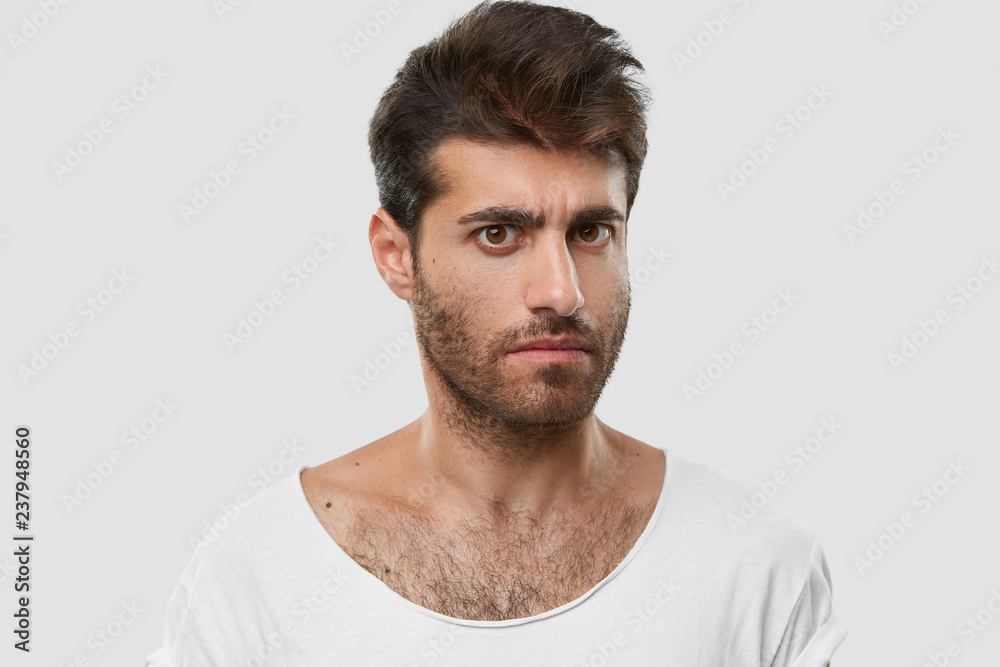 Serious young Caucasian man with dark thick stubble, trendy haircut, casual  clothes, models against white background, frowns eyebrows, being angry with  someone. Masculinity and lifestyle concept Stock Photo | Adobe Stock