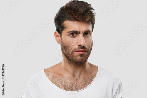 Serious young Caucasian man with dark thick stubble, trendy haircut, casual clothes, models against white background, frowns eyebrows, being angry with someone. Masculinity and lifestyle concept
