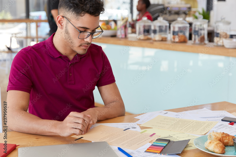 Naklejka premium Image of focused male student prepares report in finances, looks attentively at papers, eats delicious croissants, poses over cafe interior with free space for your promotion. Freelance work