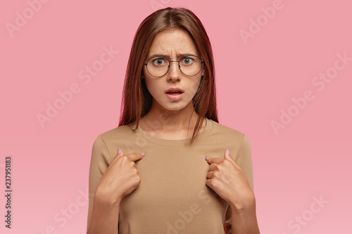 Image of indignant European woman with dark hair, points at chest, expresses negative emotions, asks why she should do housework, wears spectacles, isolated over pink background. Do you ask me?