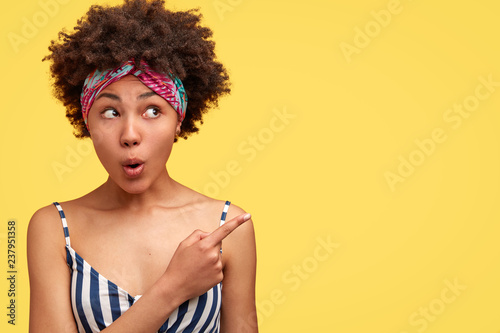 Amazed dark skinned young woman has curly hairstyle  opens mouth with surprisement  indicates at upper right corner  demonstrates something unbelievable  wears fashionable clothes  models indoor