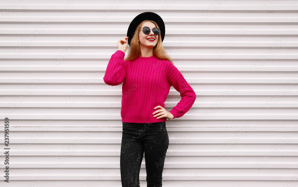 Stylish happy smiling woman in colorful pink knitted sweater, black round hat on white wall background