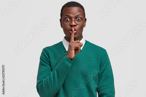 Surprised attractive black man keeps index finger over mouth, demonstrates silence sign, asks not tell his secret, wears green jumper, isolated over white background. People and secrecy concept