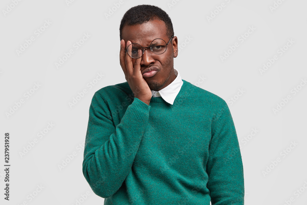 Cheereless pity dark skinned guy touches face with palm, looks in displeasure through round spectacles, purses lips, dressed in green sweater, models over white background. Negative feelings concept Cheereless pity dark skinned guy touches face with palm, looks in displeasure through round spectacles, purses lips, dressed in green sweater, models over white background. Negative feelings concept