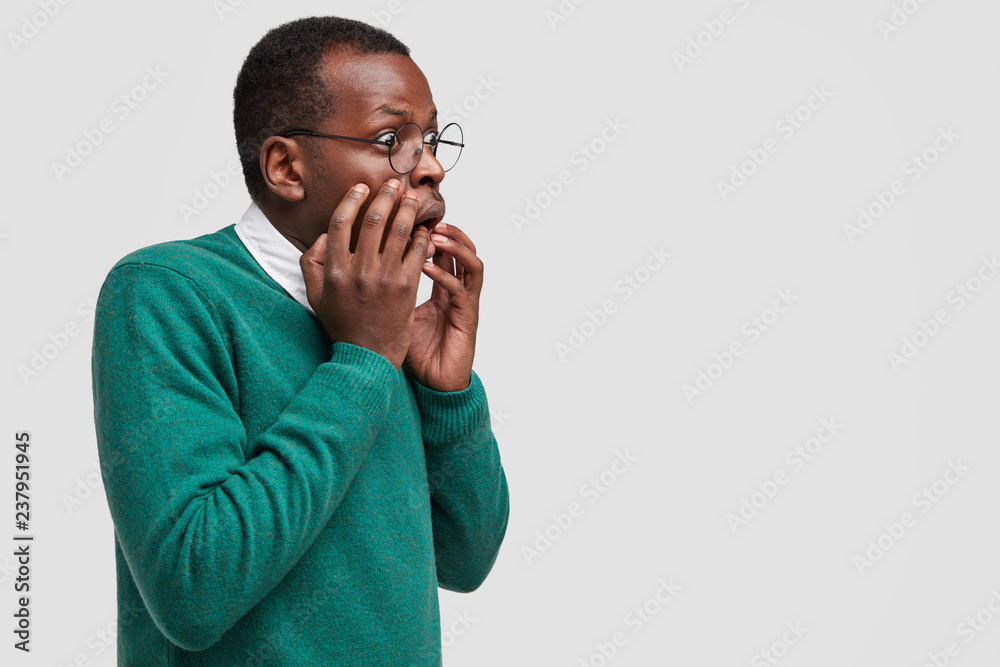 Profile shot of aghast dark skinned business owner exclaims with anger, stares aisde, poses sideways at camera, wears green sweater, isolated over white background with free space for information