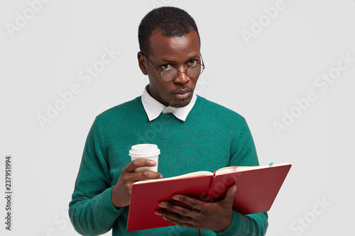 Serious dark skinned male wonk has short culry hair, holds textbook, take out coffee, looks through spectacles, wears green sweater studies indoor against white background. People and studying concept photo