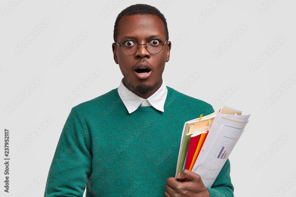 Image of stupefied male employee holds papers with financial report and diagrams, scientific literature, prepares for having business lessons, surprised by analyzed data, isolated on white wall Image of stupefied male employee holds papers with financial report and diagrams, scientific literature, prepares for having business lessons, surprised by analyzed data, isolated on white wall