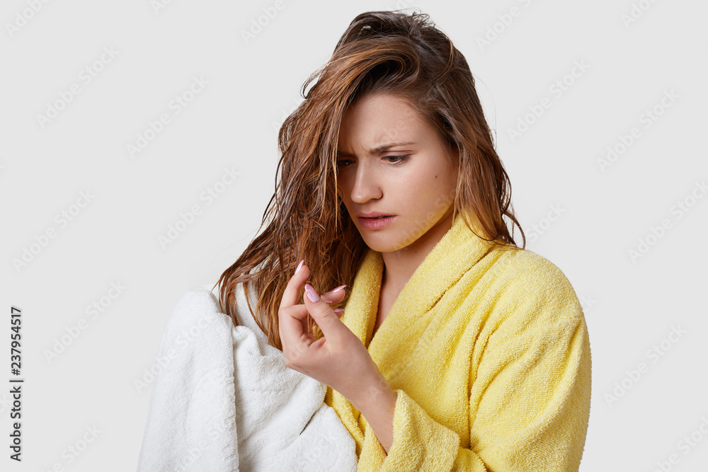Displeased young woman realizes she has damaged hair, looks stressfully at ends, being wet after taking shower, wipes with white towel, wears yellow bathrobe, isolated over white studio wall Displeased young woman realizes she has damaged hair, looks stressfully at ends, being wet after taking shower, wipes with white towel, wears yellow bathrobe, isolated over white studio wall